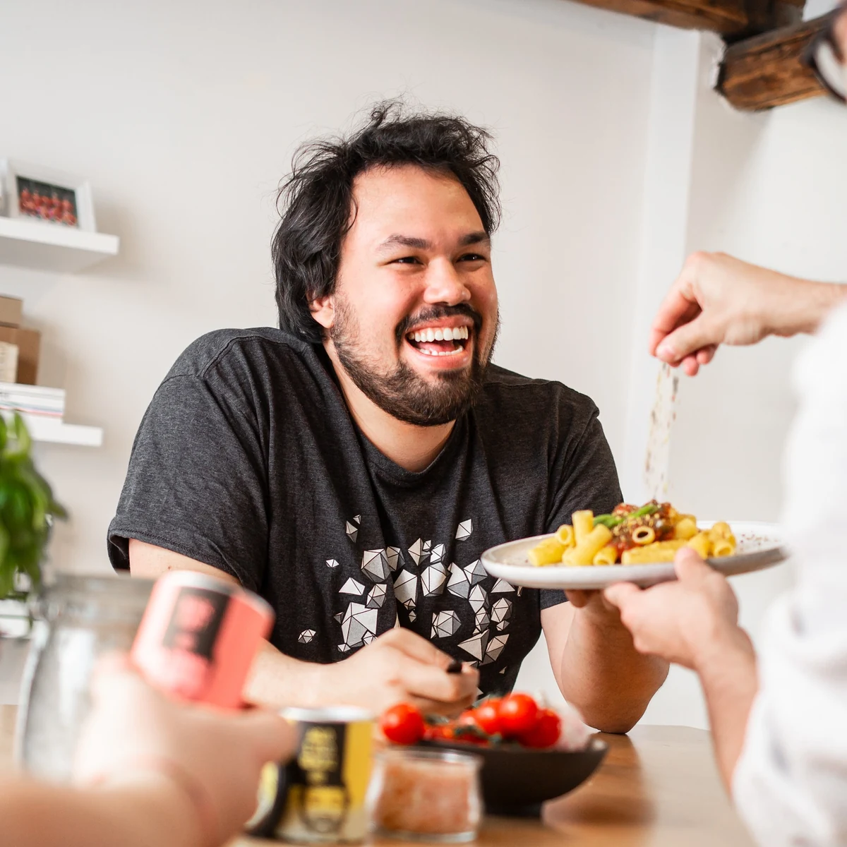 Ein lachender Mann am Tisch, während im Vordergrund ein Teller Pasta gewürzt wird.